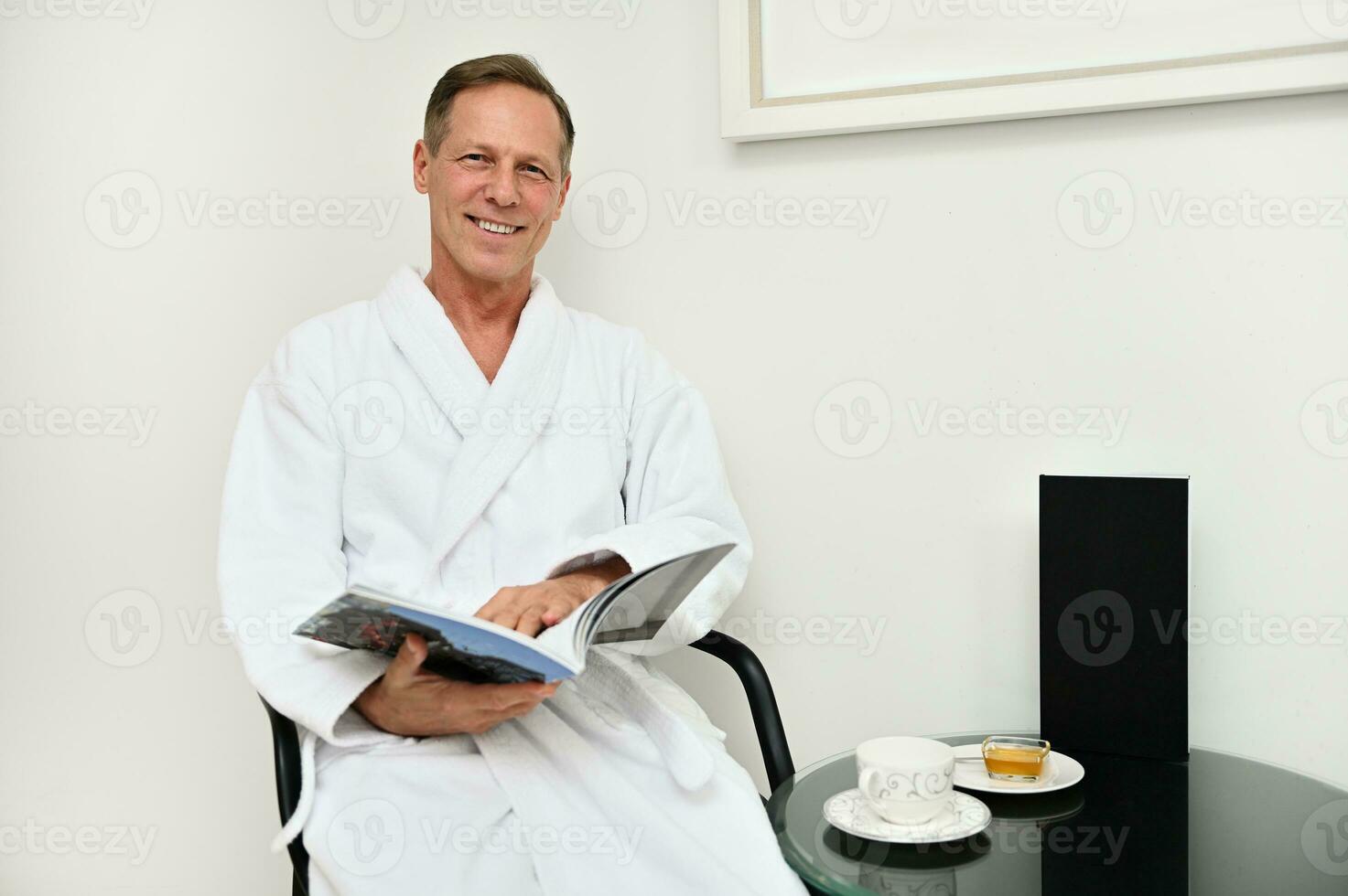 knap charmant volwassen Kaukasisch Mens in wit badstof badjas zittend Aan fauteuil met een tijdschrift in zijn handen, glimlacht toothy glimlach op zoek Bij camera, ontspannende Bij luxueus welzijn spa centrum foto