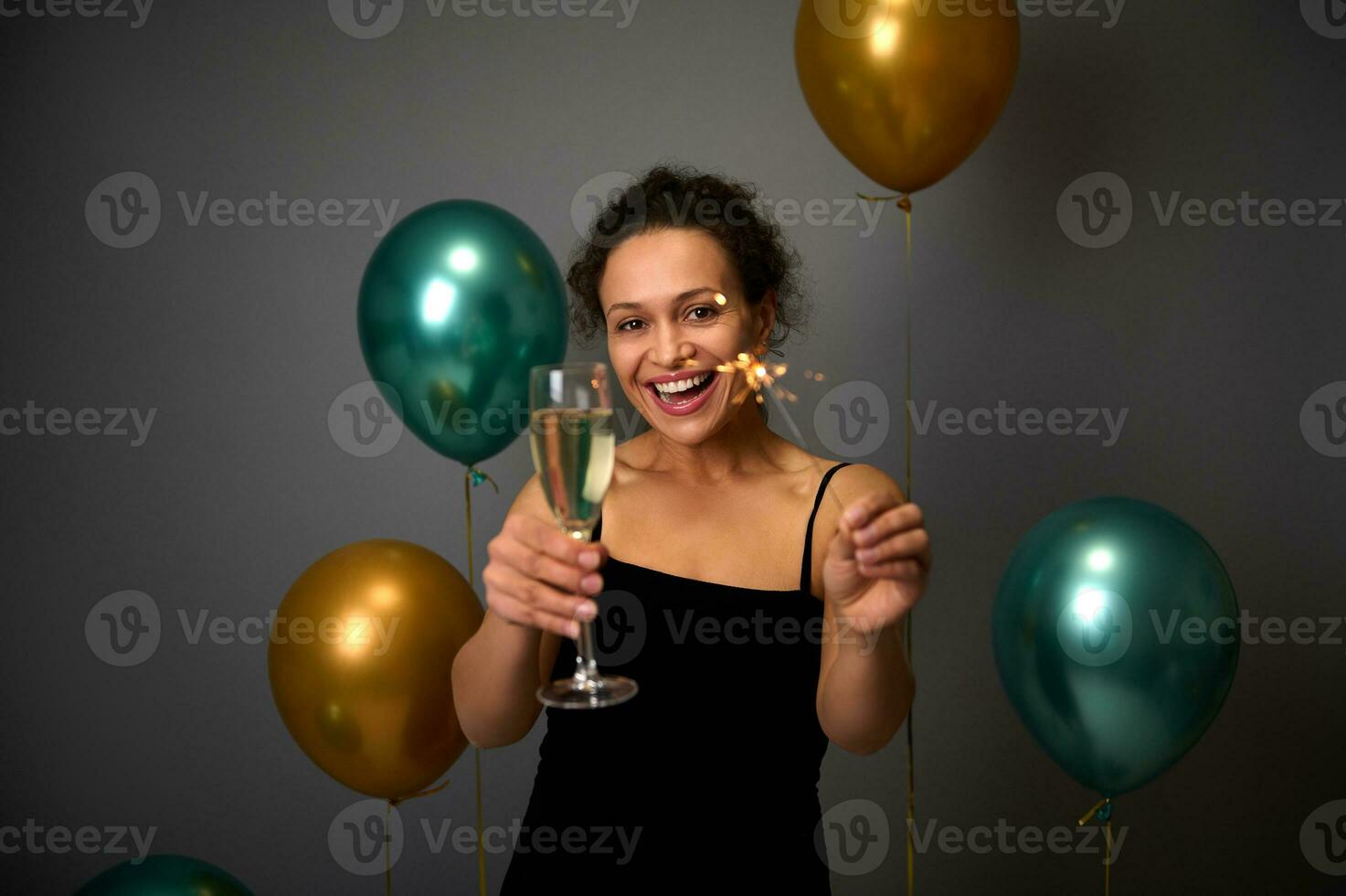 gelukkig Afrikaanse Amerikaans vrouw houdt fluit van Champagne en sterretjes, glimlacht met mooi toothy glimlach poes tegen grijs muur achtergrond met opgeblazen gouden en groen lucht ballonnen. kopiëren ruimte voor advertentie foto