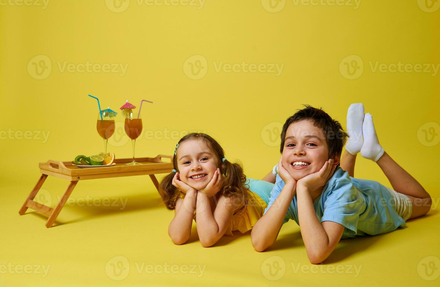 mooi kinderen aan het liegen in de buurt een bamboe dienblad met cocktails in glas versierd met paraplu. geïsoleerd Aan geel achtergrond met kopiëren ruimte foto
