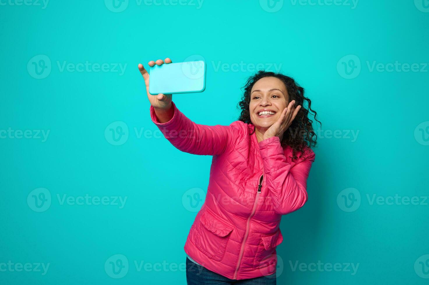 aantrekkelijk donker haar geweldig vrouw met gekruld kapsel, vervelend roze jasje, Holding hand- in de buurt haar gezicht, glimlacht toothy glimlach op zoek Bij smartphone camera, maken selfie. blauw achtergrond kopiëren ruimte foto