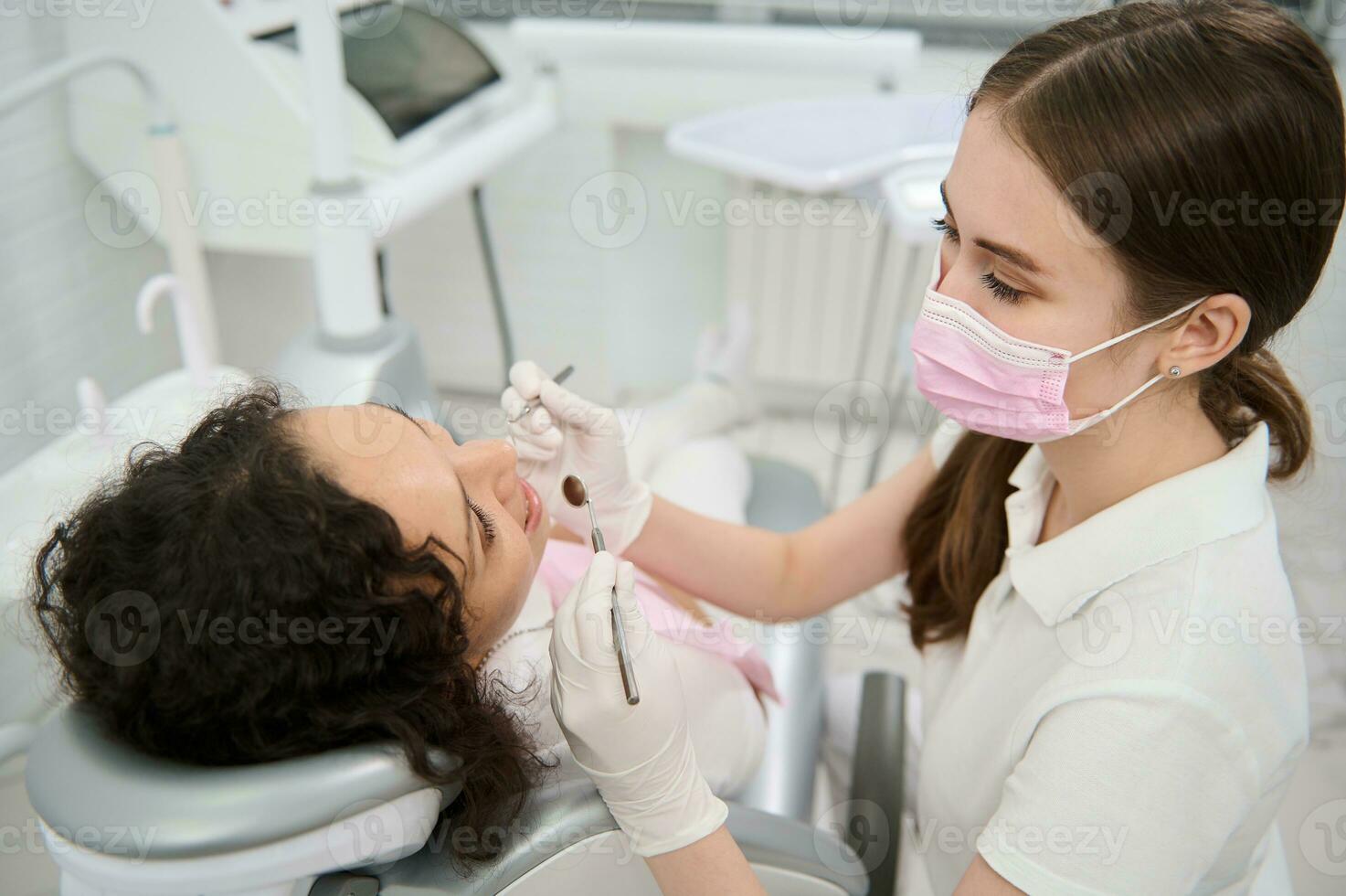 tandarts hygiënist in medisch beschermend masker onderzoeken de mondeling holte van een vrouw geduldig met steriel roestvrij staal tandheelkundig spiegel en sonde in tandheelkunde kliniek gedurende gepland medisch controle foto