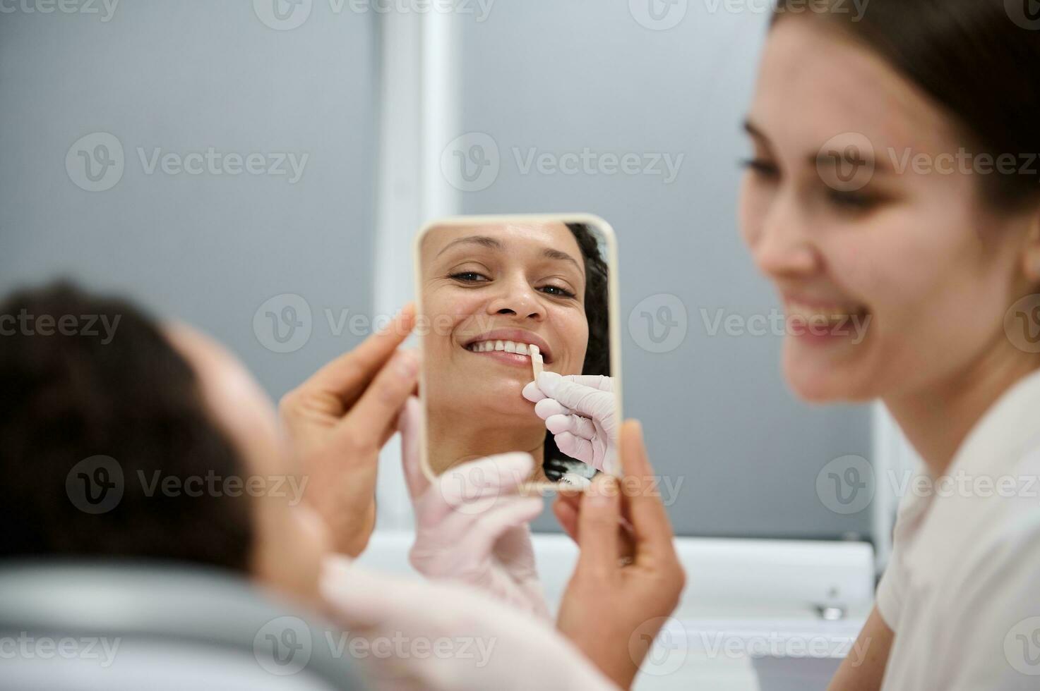 detailopname van midden- volwassen vrouw Holding in de buurt haar tanden een kleur tabel voor bleken tanden en vergelijken de witheid van tanden met tandheelkundig steekproef, glimlachen toothy glimlach op zoek Bij haar spiegel reflectie foto