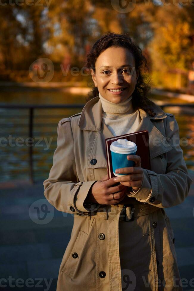 portret van een vrolijk Afrikaanse vrouw met meenemen kop van koffie en een hand- boek glimlachen op zoek Bij camera Aan de herfst park meer achtergrond met vallend bladeren. herfst- buitenshuis vrije tijd. recreatie foto