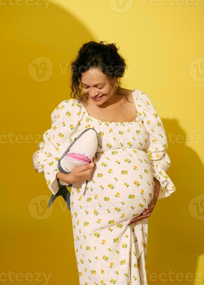 vrolijk zwanger vrouw lachend, Holding pluche haai speelgoed, geïsoleerd over- geel studio achtergrond foto