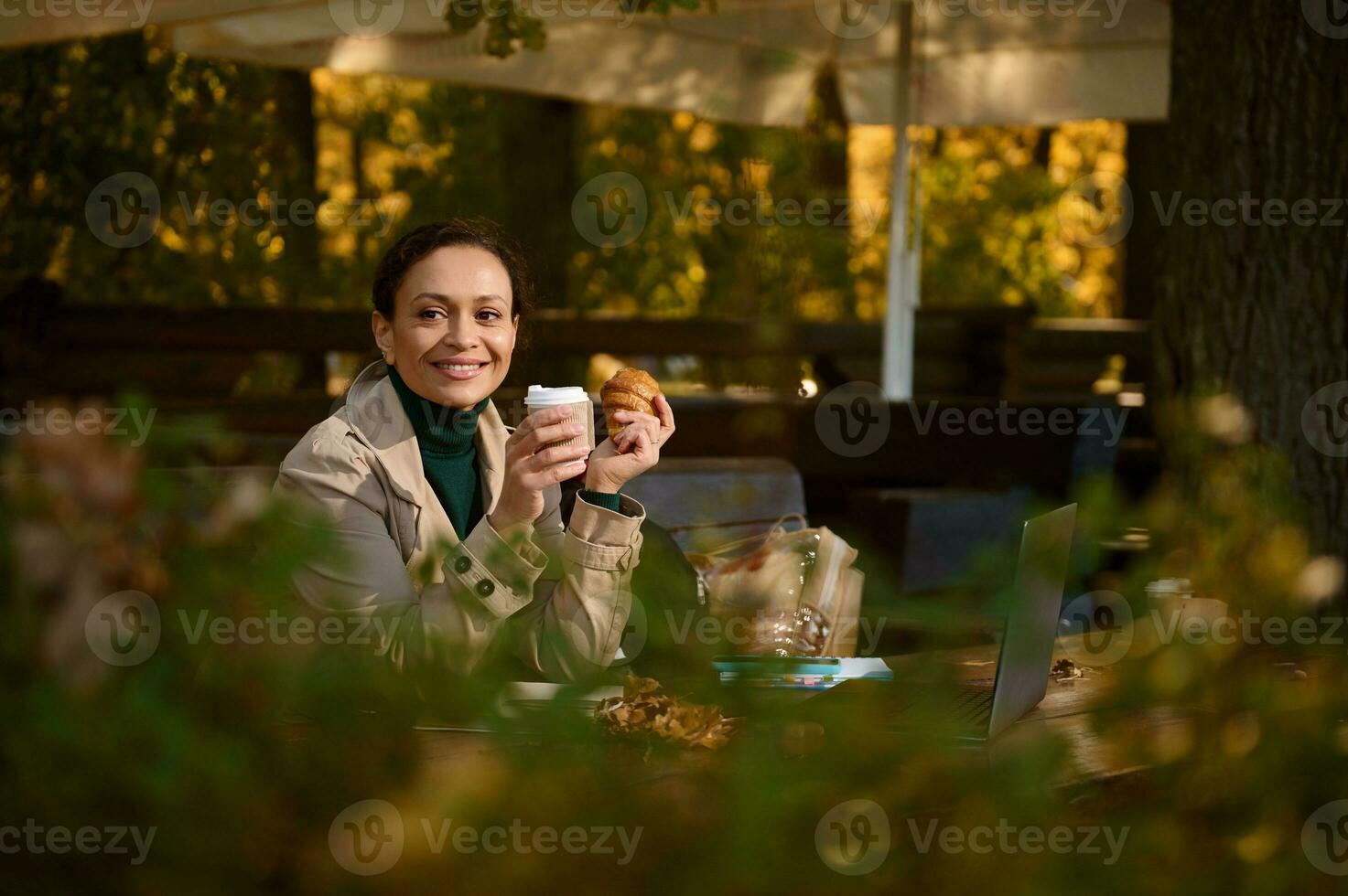 aantrekkelijk donker haar bedrijf vrouw vervelend beige jas hebben een koffie breken in een buitenshuis terras van een houten cafetaria in eik bosje Aan mooi zonnig herfst dag. bezig vrouw resting na werk foto