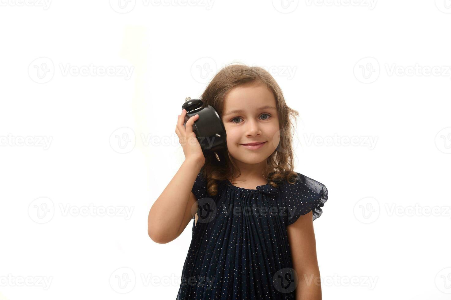een mooi 4 jaren oud weinig meisje in een donker blauw jurk houdt een alarm klok in de buurt haar oor en luistert aandachtig naar de geluid, glimlacht op zoek Bij camera, geïsoleerd Aan wit achtergrond met kopiëren ruimte foto