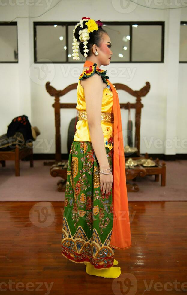 een traditioneel Indonesisch danser staand in een geel kostuum en een oranje sjaal hangende naar beneden haar lichaam foto