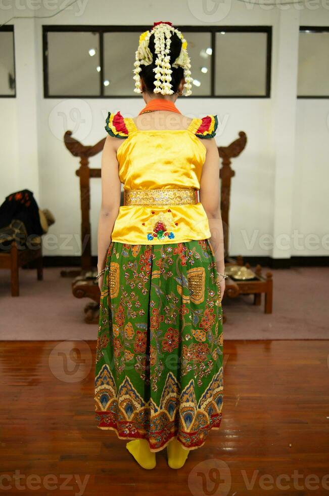 een traditioneel Indonesisch danser staand in een geel kostuum en een oranje sjaal hangende naar beneden haar lichaam foto