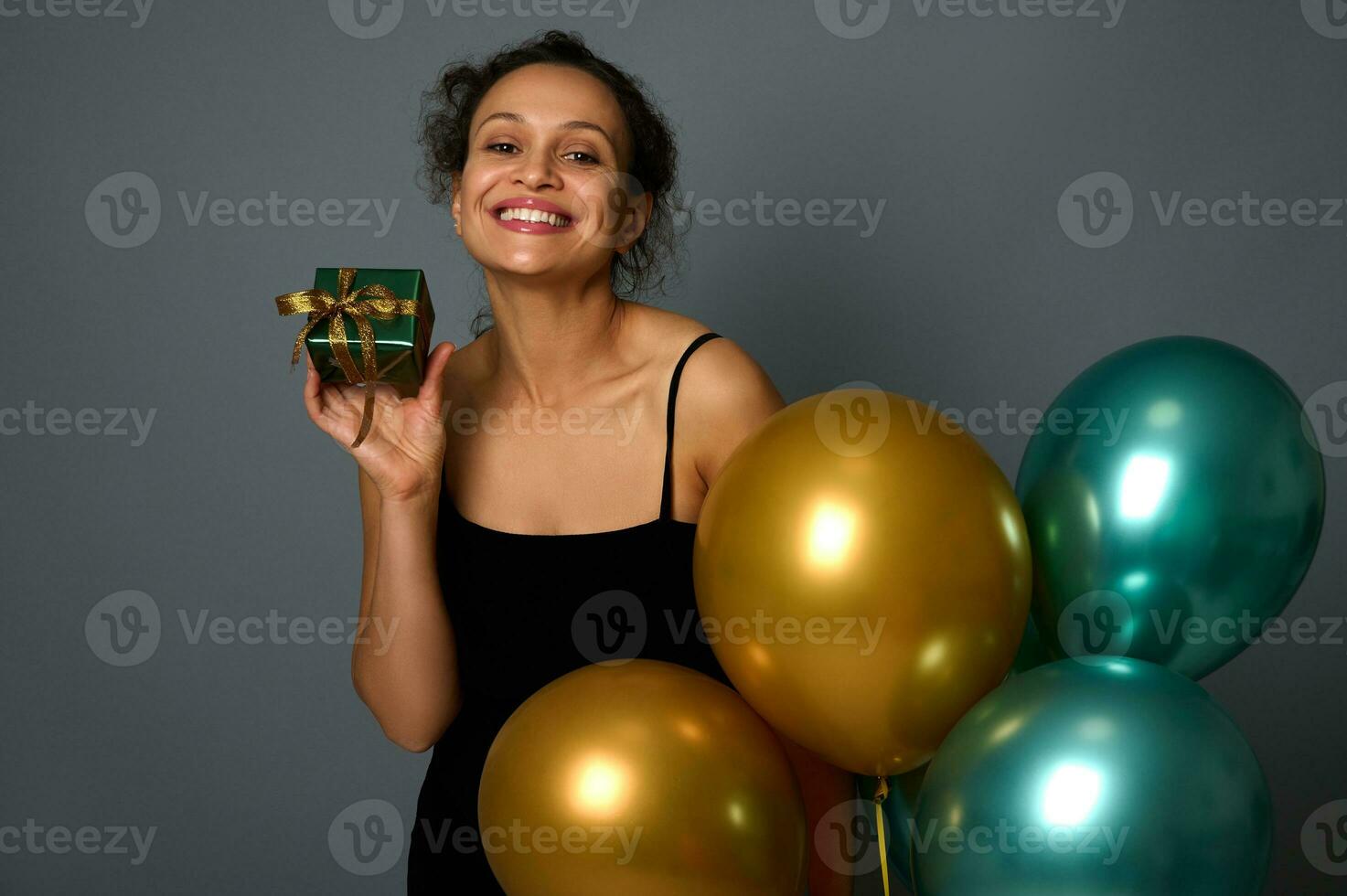 detailopname van mooi vrouw met mooi glimlach poseren met een Kerstmis geschenk in glimmend omhulsel papier en gouden boog, Holding feestelijk mooi opgeblazen lucht ballonnen Aan grijs achtergrond met kopiëren advertentie ruimte foto