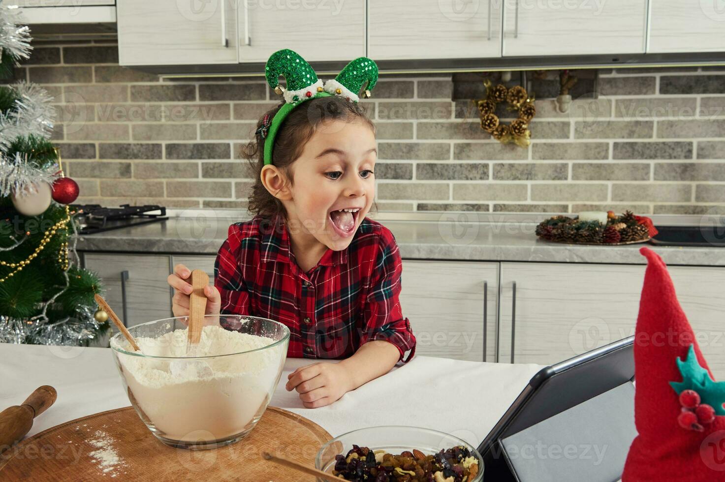 aanbiddelijk meisje, weinig kind vlogger opname video terwijl staand door tafel en Koken Kerstmis gebakje, menging ingrediënten in de kom en op zoek Bij scherm toezicht houden op van een digitaal tablet foto