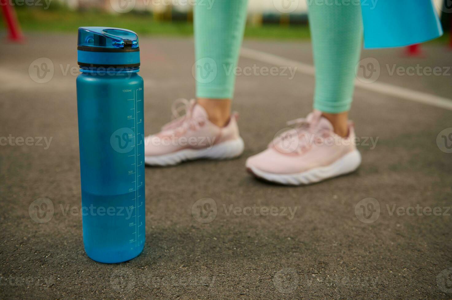 detailopname van een blauw plastic fles van water Aan de asfalt van sport- grond De volgende naar sportief vrouw voeten. Gezondheid en lichaam zorg concept foto