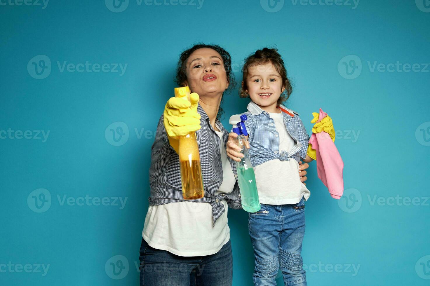 mooi jong vrouw, huisvrouw regisseert schoonmaak sprays net zo als het schieten van een pistool, staand De volgende naar haar weinig aanbiddelijk dochter . huis werk en klusjes concept foto