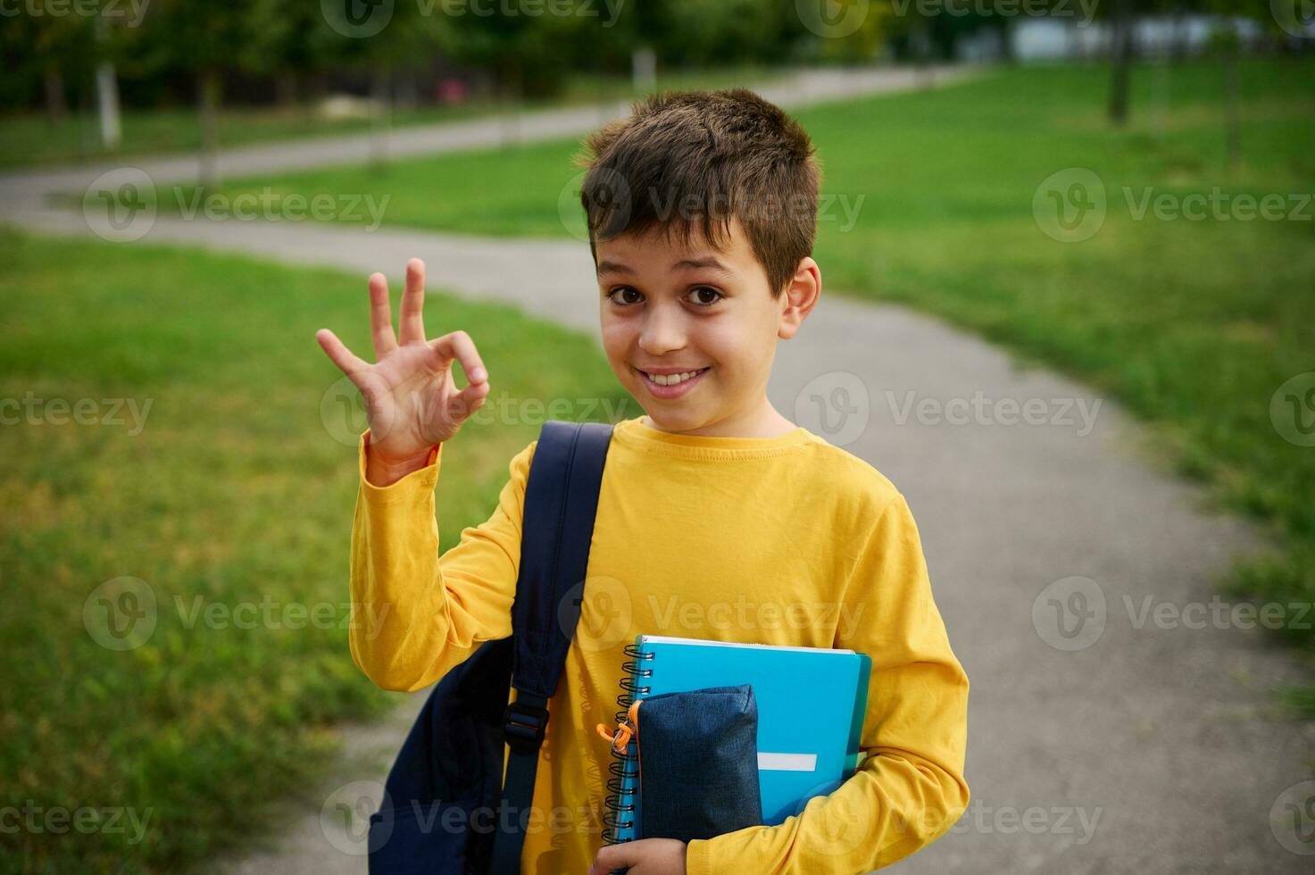 knap aanbiddelijk schooljongen tonen zingen OK met zijn hand, staand met rugzak en school- benodigdheden in de stad park foto