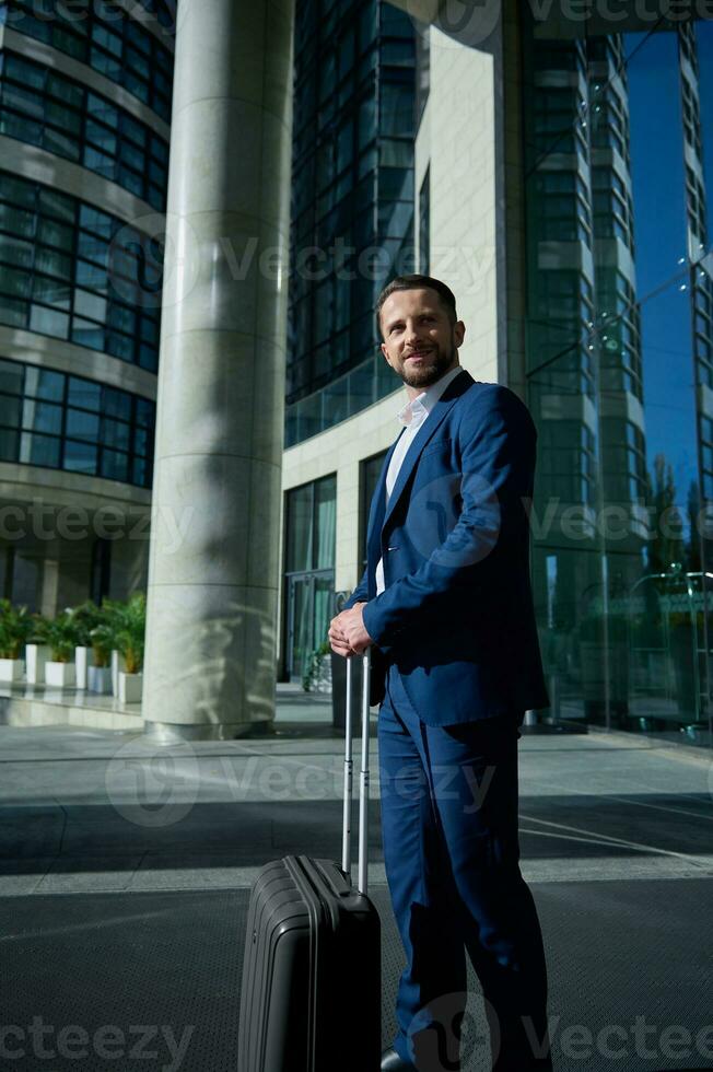 zelfverzekerd aantrekkelijk geslaagd zakenman ontwikkelaar ondernemer in bedrijf pak en bagage glimlacht op zoek terzijde staand buitenshuis Aan een zakelijke hoogbouw gebouw glas Ingang achtergrond foto