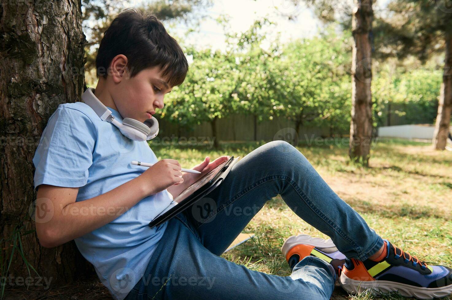 spaans tiener jongen genieten van zijn creatief hobby, tekening schetsen Aan een digitaal tablet, gebruik makend van een stylus pen, zittend in park foto