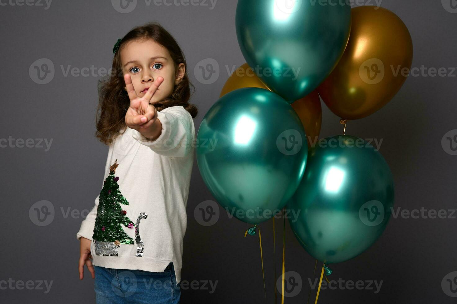 weinig meisje in Kerstmis trui staat in de buurt mooi glimmend groen metalen en gouden lucht ballen en shows een vrede teken, gebaren met haar vinger, geïsoleerd over- grijs achtergrond met kopiëren ruimte voor advertentie foto