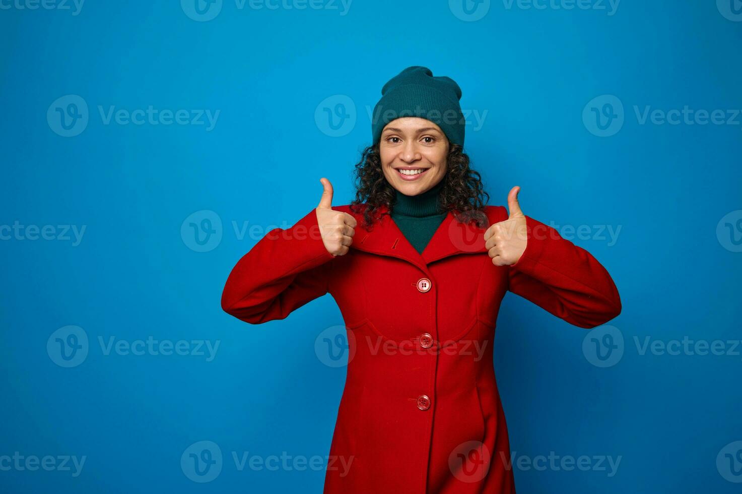 charmant geweldig aantrekkelijk mooi vrouw in helder rood jas en groen hoed glimlachen met vrolijk toothy glimlach, op zoek Bij camera en tonen duimen omhoog, poseren tegen blauw achtergrond met kopiëren ruimte foto