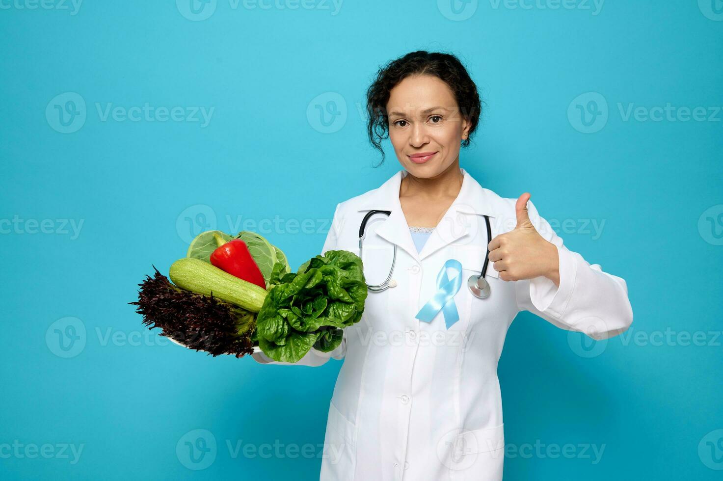 vrouw dokter in wit medisch gewaad, vervelend blauw satijn lint, symbolisch boog kleur verhogen bewustzijn in diabetes dag, Holding een bord met rauw veganistisch gezond maaltijd en shows duim omhoog op zoek Bij camera foto