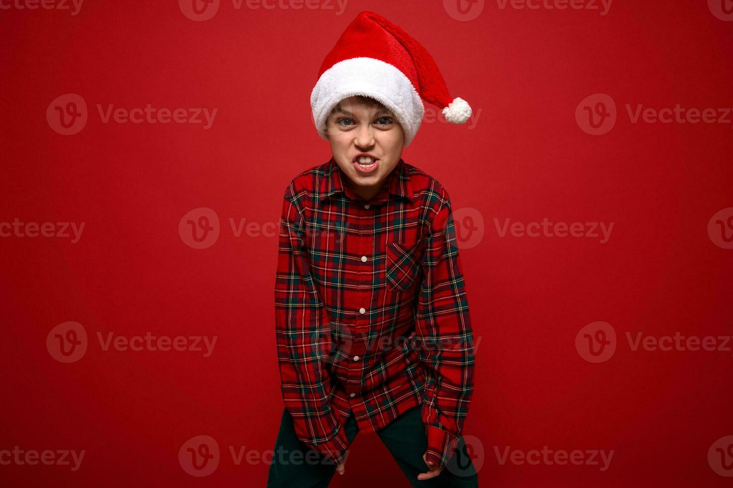 grappig preadolescent jongen in plaid overhemd en de kerstman claus hoed grimassen op zoek Bij camera poseren tegen rood gekleurde achtergrond met kopiëren ruimte voor Kerstmis en nieuw jaar advertentie foto