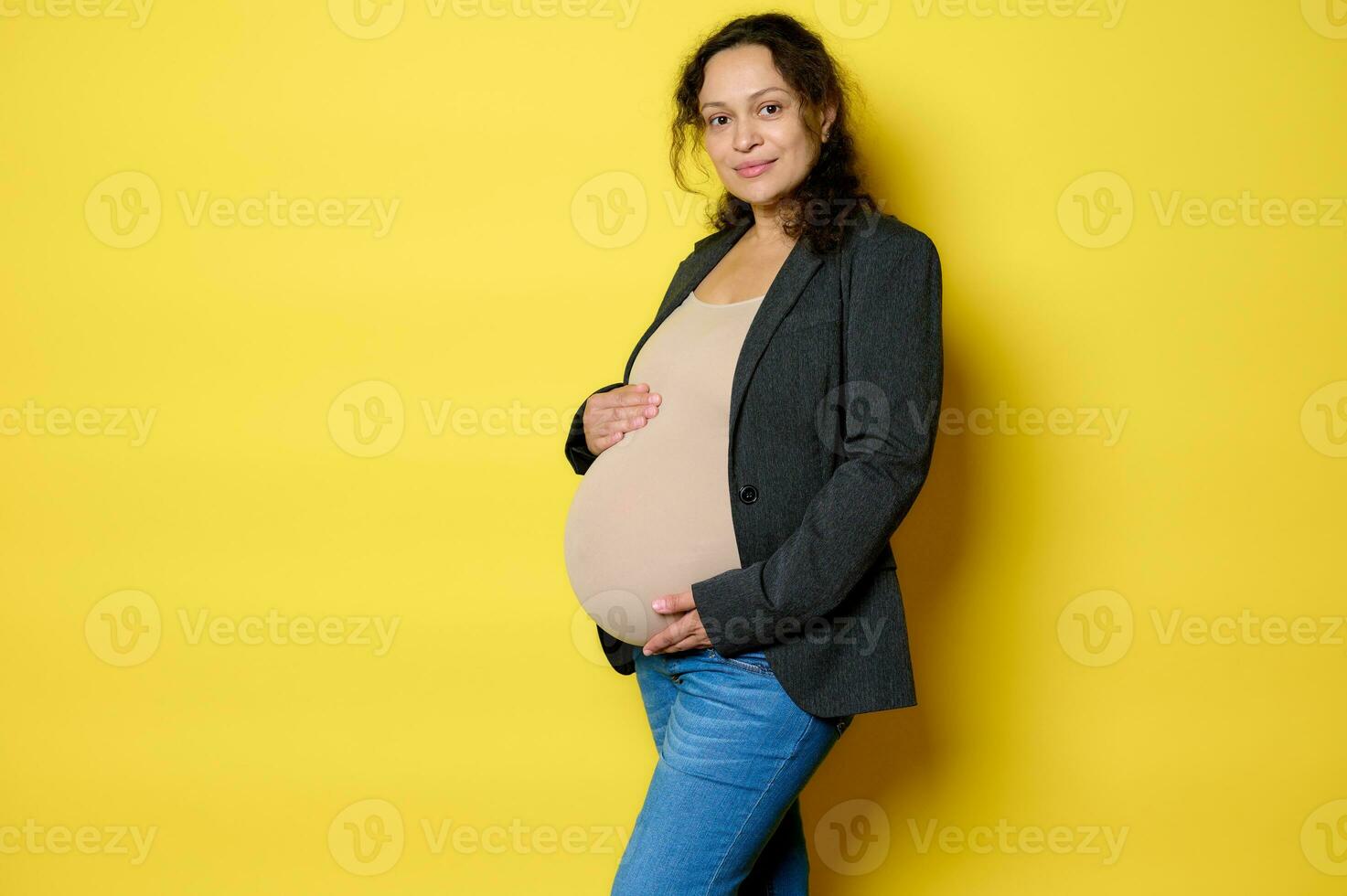 aantrekkelijk latina zwanger vrouw in elegant blazer, bodysuit en blauw jeans, glimlacht Bij camera, poses met handen Aan buik foto