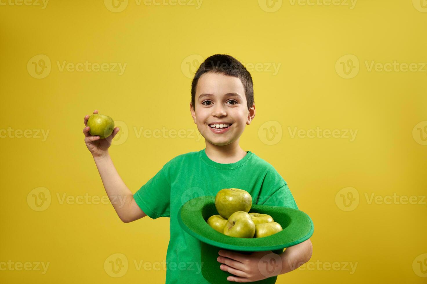 grappig weinig jongen staat geïsoleerd Aan geel achtergrond en houdt een Iers elf van Ierse folklore hoed vol van appels in een hand- en een groen appel in de ander. heilige Patrick dag foto