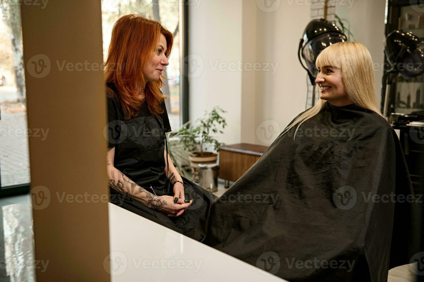 modieus mooi roodharig vrouw in zwart professioneel uniform, haar- stilist met elegant lichaam tatoeages Aan armen pratend met een haar- salon klant zittend in kapper stoel in voorkant van een spiegel foto