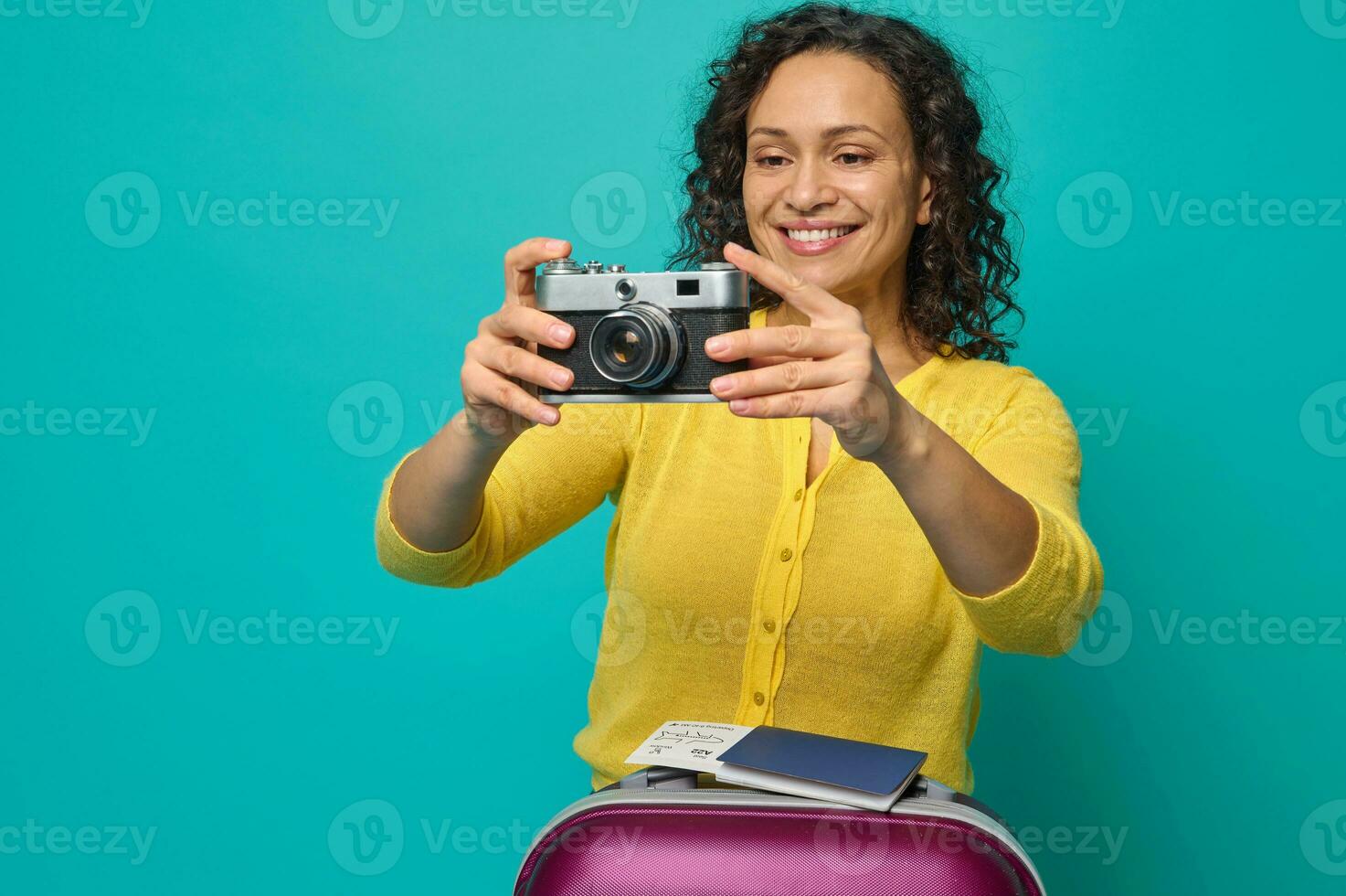 aantrekkelijk midden volwassen spaans vrouw in helder geel trui fotograferen met een wijnoogst camera, glimlachen toothy glimlach poseren met koffer, instappen voorbij gaan aan en ticket Aan blauw achtergrond. kopiëren ruimte foto