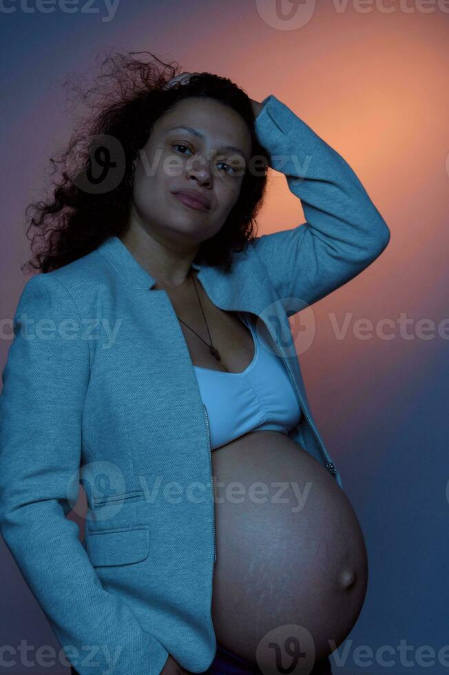 mooi multi-etnisch zwanger vrouw, op zoek Bij camera, poseren met naakt buik Aan studio achtergrond met neon lichten foto