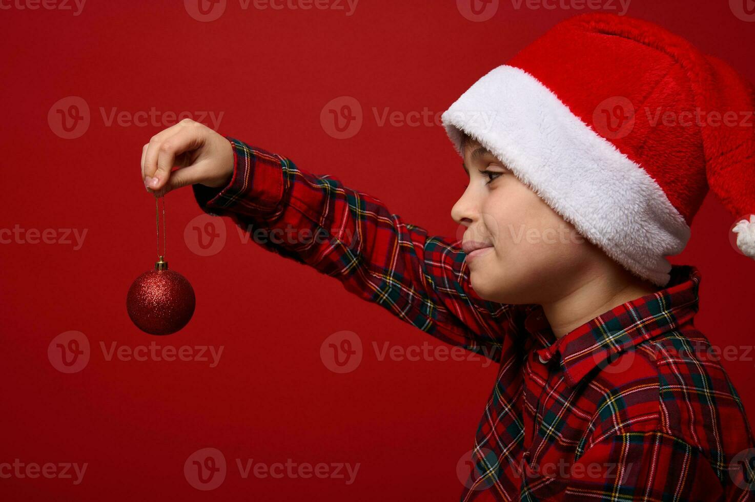 kant detailopname portret van een aanbiddelijk kind jongen in een de kerstman hoed Holding een rood glimmend bolvormig speelgoed- van de Kerstmis boom in voorkant van hem, poseren Aan een gekleurde achtergrond met kopiëren ruimte voor nieuw jaar advertentie foto