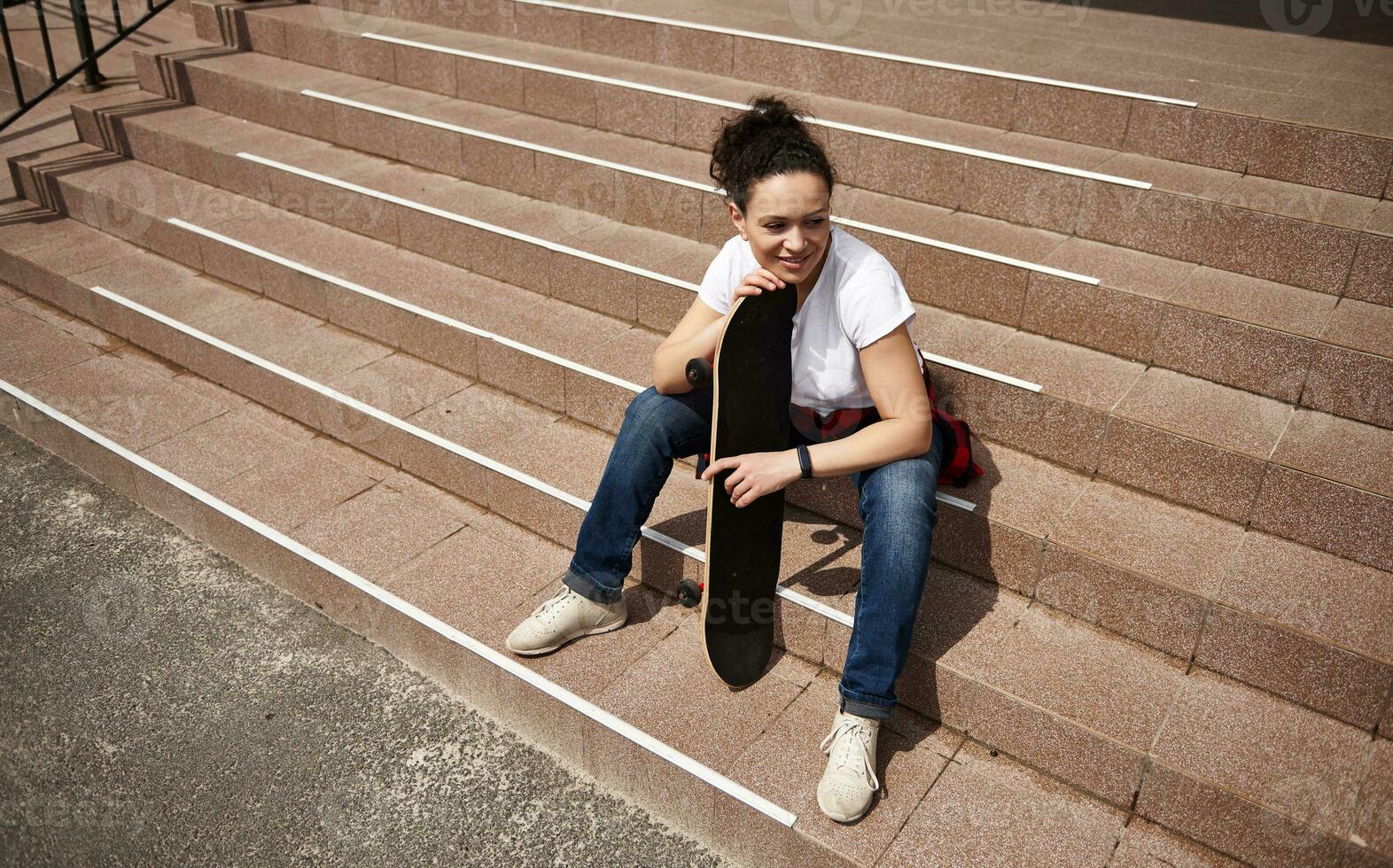 hoog hoek visie van een gelukkig glimlachen vrouw met skateboard zittend Aan stappen buitenshuis foto