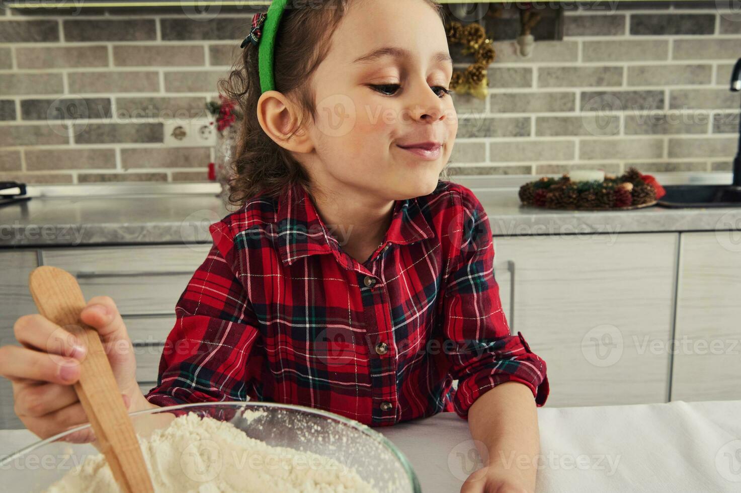 detailopname van een mooi positief glimlachen Europese weinig meisje in rood en groen geruit jurk menging ingrediënten in een glas kom met een houten lepel, Koken Kerstmis gebakje Bij huis keuken foto