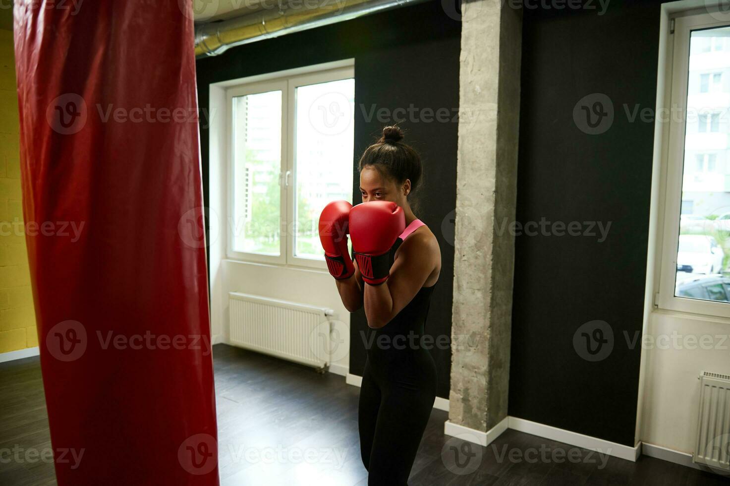 geschiktheid vrouw bokser in nauwsluitend zwart sportkleding en rood boksen handschoenen opleiding in een boksen Sportschool, maken een Rechtdoor stempel raken een ponsen zak gedurende een binnen- training foto