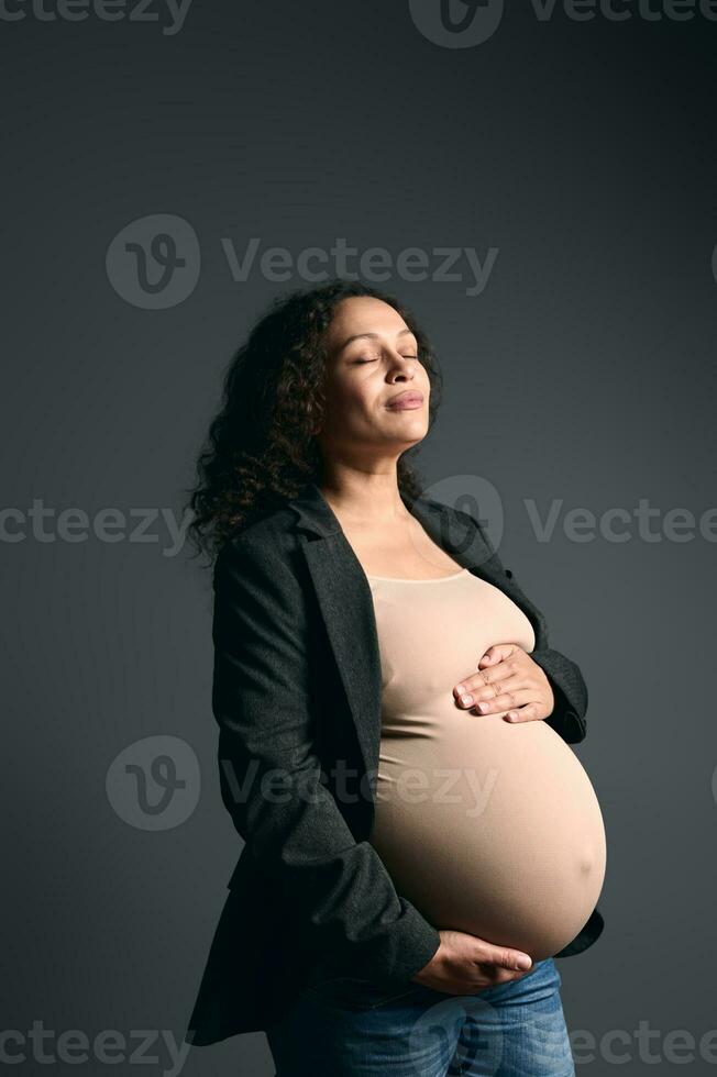 verrukkelijk sereen zwanger vrouw zetten handen Aan haar groot buik, poseren met haar ogen Gesloten over- grijs studio achtergrond foto