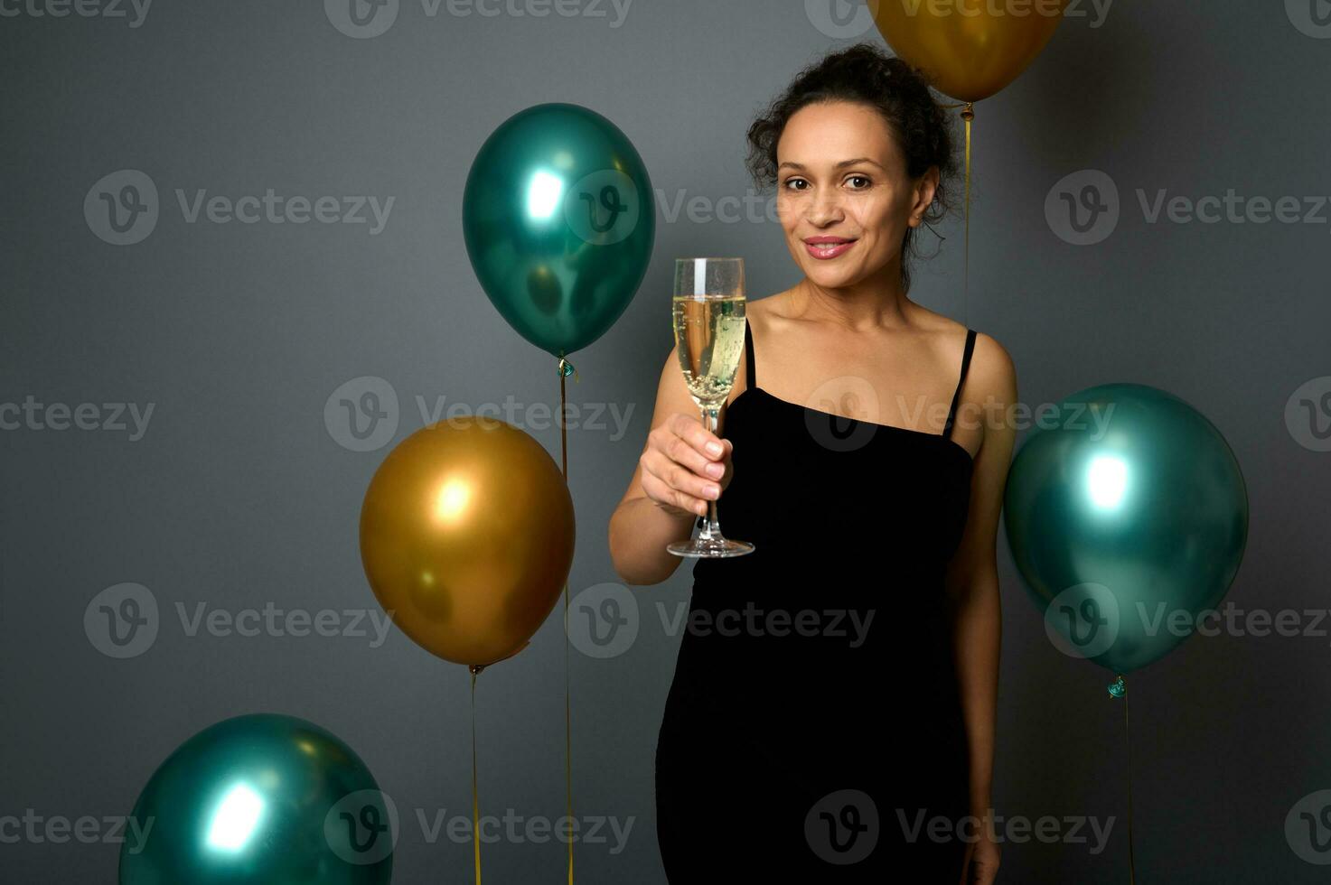 vrolijk Kerstmis en gelukkig nieuw jaar. promotionele schot van mooi gemengd ras vrouw in avond jurk met glas van Champagne poseren tegen grijs achtergrond met glimmend goud en groen metalen ballonnen foto