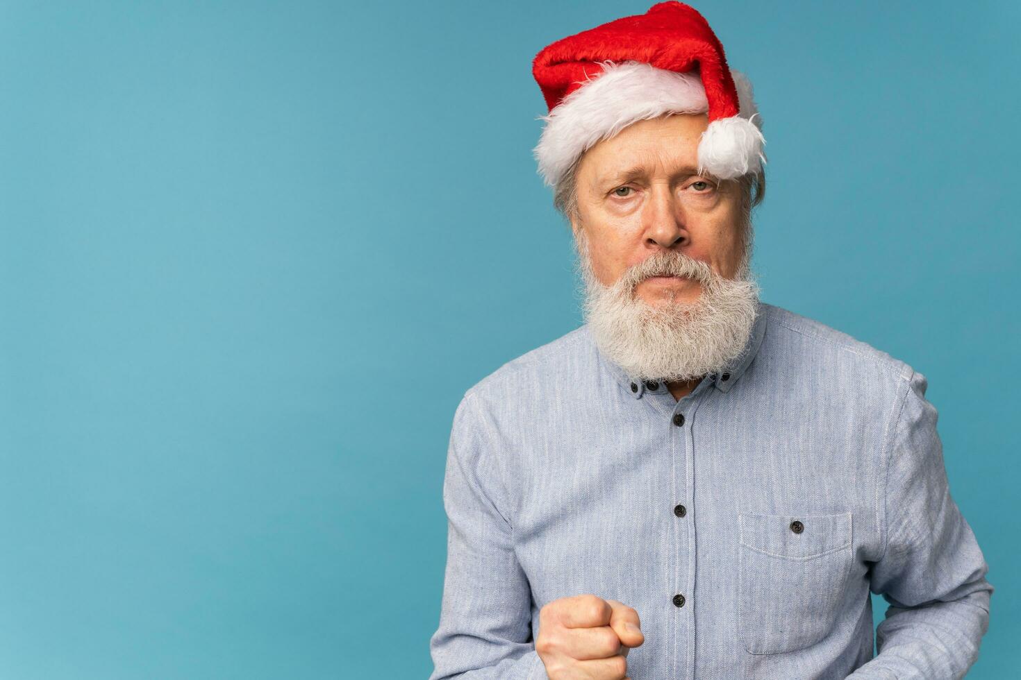 studio portret van wit baard oud Mens in de kerstman hoed tonen vuist en op zoek Bij camera met boos uitdrukking kopiëren ruimte - emotie en slecht Kerstmis humeur concept foto