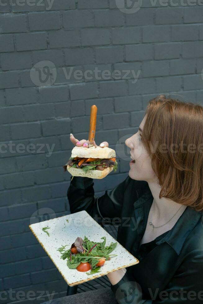 een jong vrouw eet een hamburger met vlees Aan een zomer terras foto