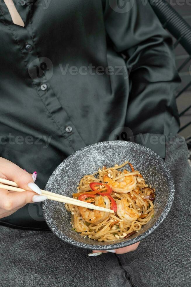 udon noedels met garnaal en gebakken groenten in een kom in vrouw handen met eetstokjes foto