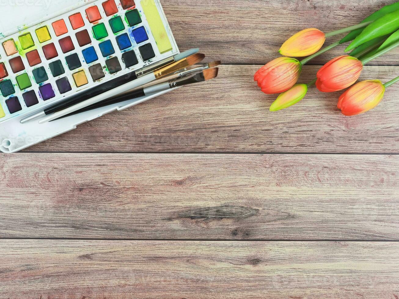 artiest werkruimte. tekening hulpmiddelen, stationair benodigdheden, waterverf verven, borstels en rood geel tulp bloemen Aan houten tafel achtergrond. foto