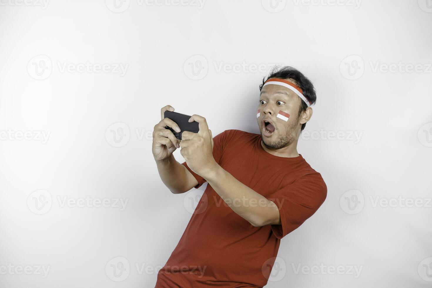 een portret van een geschokt Aziatisch Mens vervelend hoofdband, Holding zijn telefoon terwijl zijn mond breed open, geïsoleerd door wit achtergrond. Indonesië's onafhankelijkheid dag concept foto