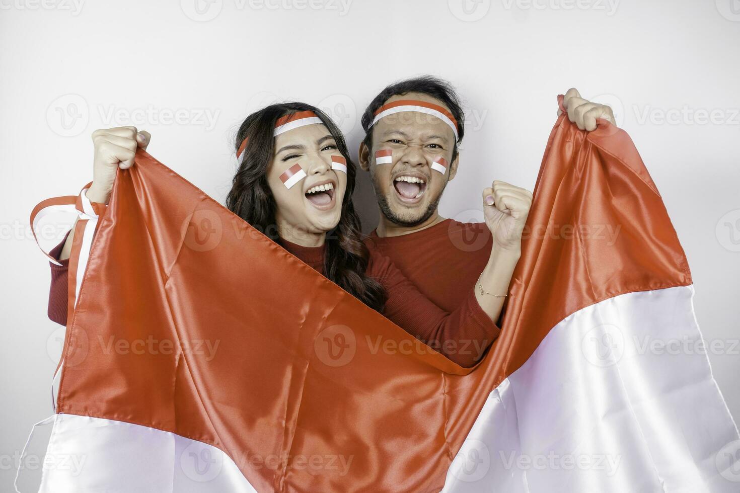 een jong Aziatisch paar met een gelukkig geslaagd uitdrukking vervelend rood top en hoofdband terwijl Holding Indonesië's vlag, geïsoleerd door wit achtergrond. Indonesië's onafhankelijkheid dag concept. foto