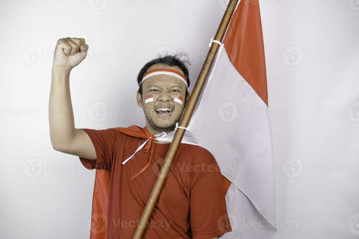 een jong Aziatisch Mens met een gelukkig geslaagd uitdrukking vervelend rood top en hoofdband terwijl Holding Indonesië's vlag, geïsoleerd door wit achtergrond. Indonesië's onafhankelijkheid dag concept. foto