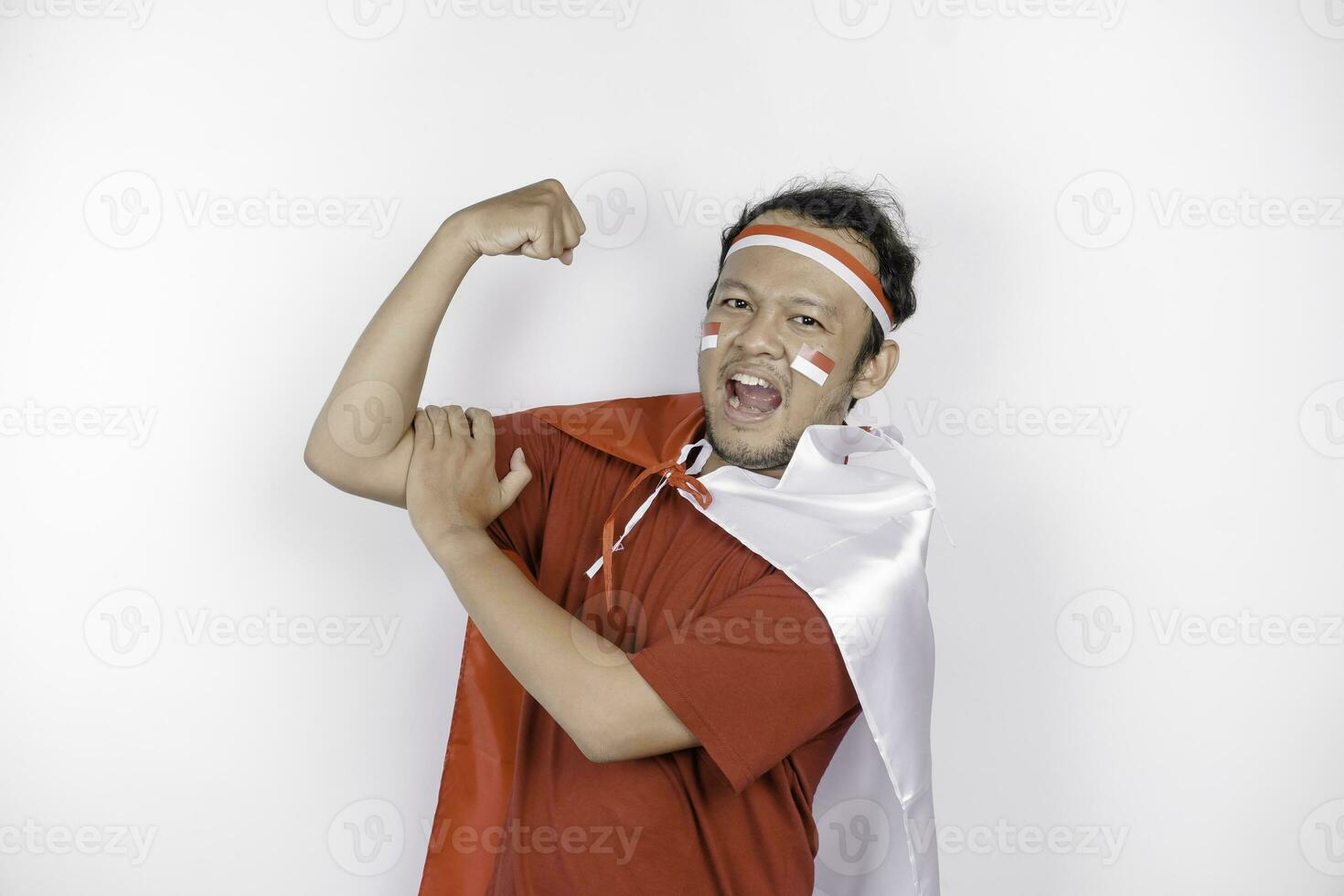 opgewonden Aziatisch Mens vervelend een rood bovenkant, vlag kaap en hoofdband, tonen sterk gebaar door hijs- zijn armen en spieren glimlachen trots. Indonesië's onafhankelijkheid dag concept. foto