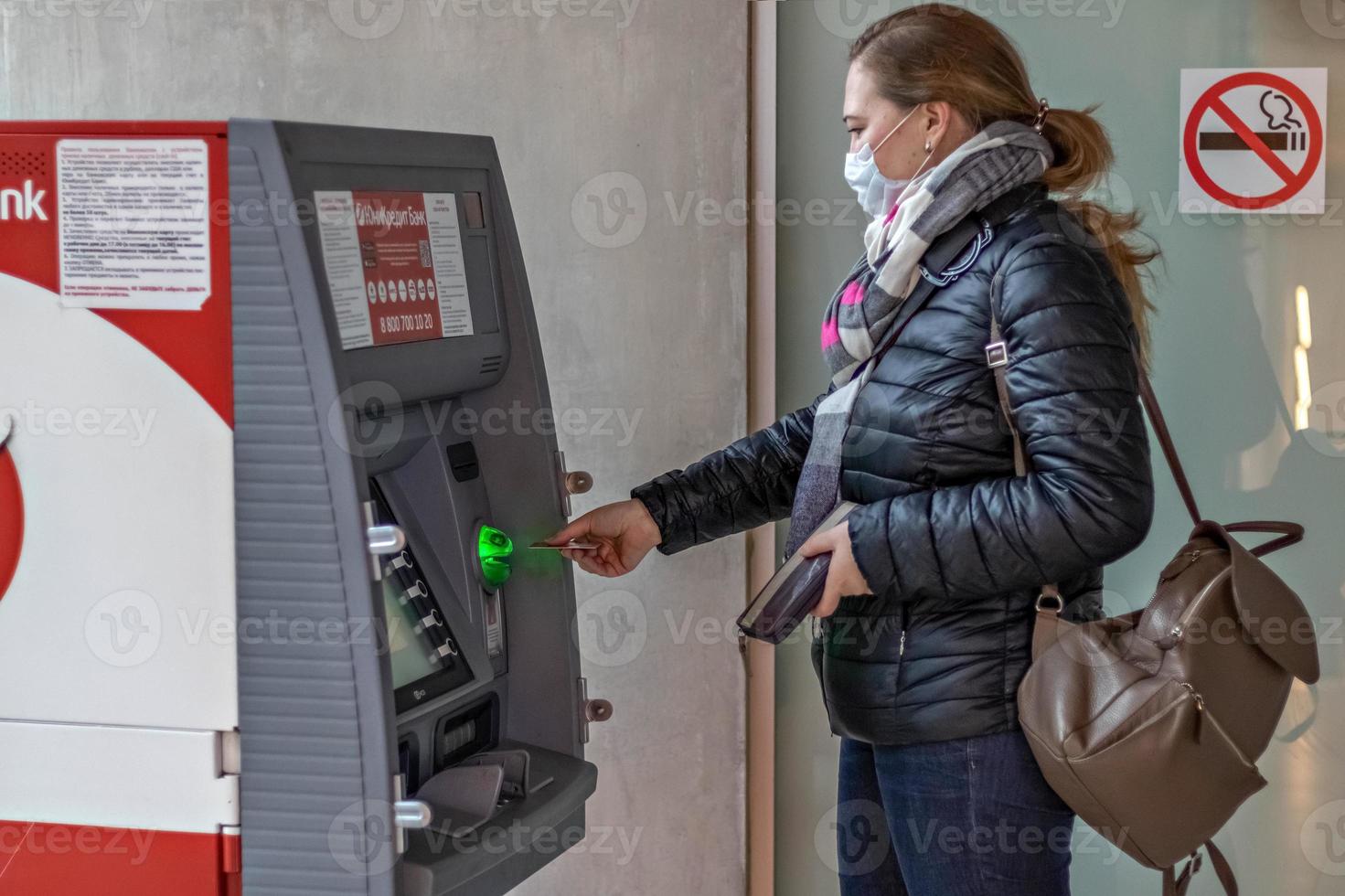 een vrouw die een beschermend medisch masker op haar gezicht draagt, steekt een creditcard uit een geldautomaat foto
