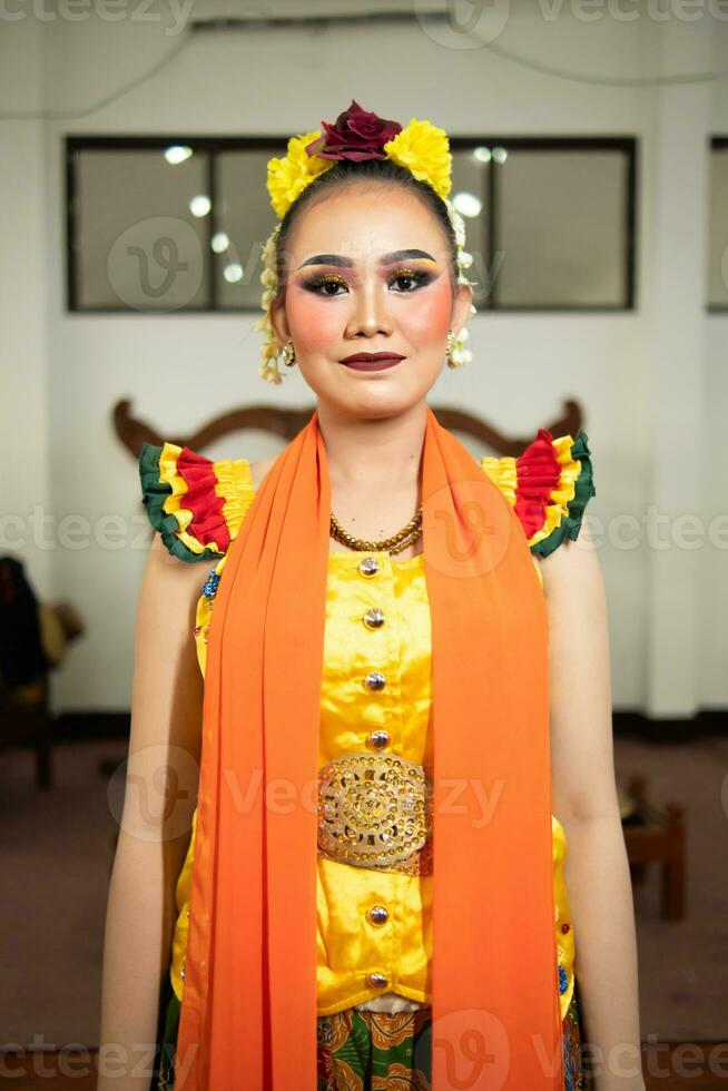 een traditioneel Indonesisch danser staand in een geel kostuum en een oranje sjaal hangende naar beneden haar lichaam foto