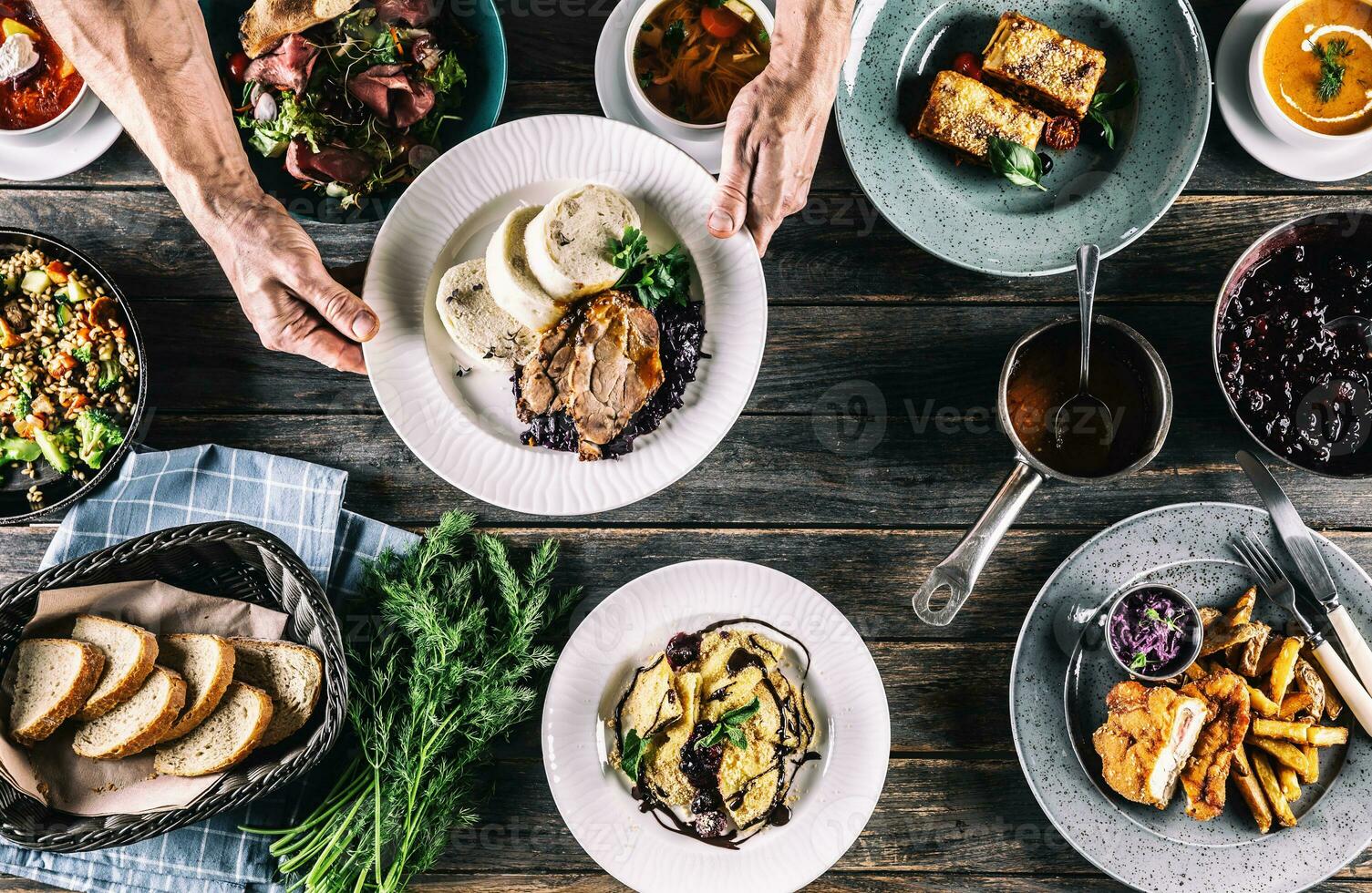 chef hand- plukken omhoog een bord met maaltijd van een top visie van een tafel vol van assortiment maaltijden , inclusief soep, brood, salade, knoedels, schnizel, pasta lasagne en kruiden foto