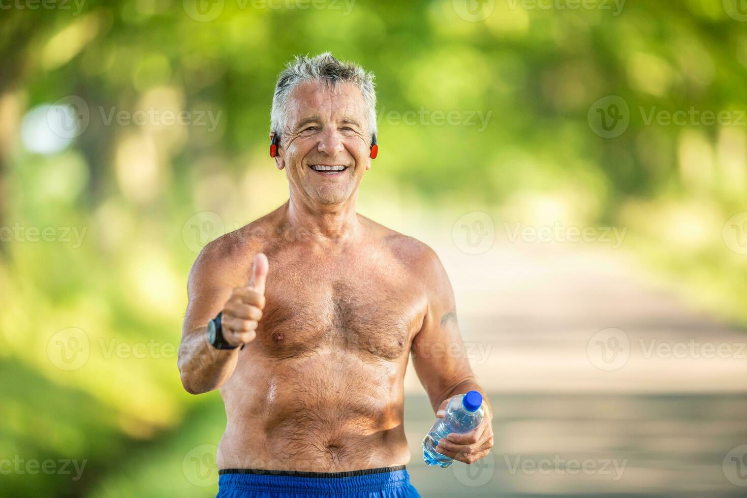 grijs haar ouder Mens shows duimen omhoog terwijl Holding gebotteld water en vervelend Nee t-shirt na een training in de natuur foto