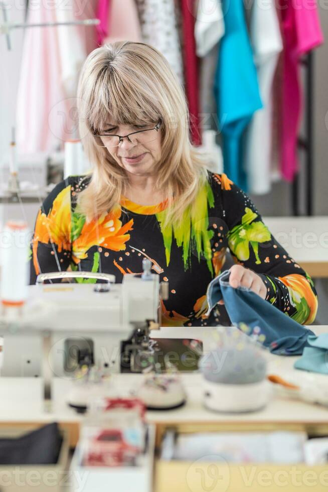 ouder vrouw zit achter de naaien machine maken kleren in de werkplaats foto