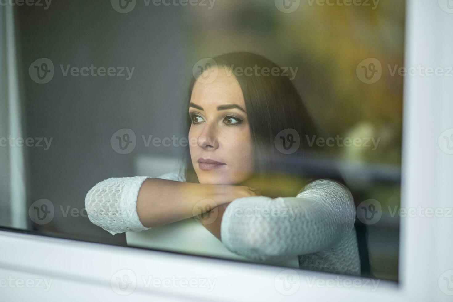 verdrietig mooi vrouw zit Aan een stoel achter de venster op zoek buiten foto