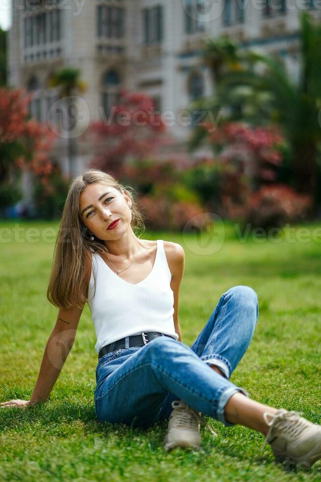 mooi jong vrouw vervelend tank top en jeans zittend Aan gras gazon. groen gazon in voorkant van oud gebouw. zonnig dag in park, groen gras en bomen in achtergrond. kalmte meisje genieten ontspannende in stad. foto