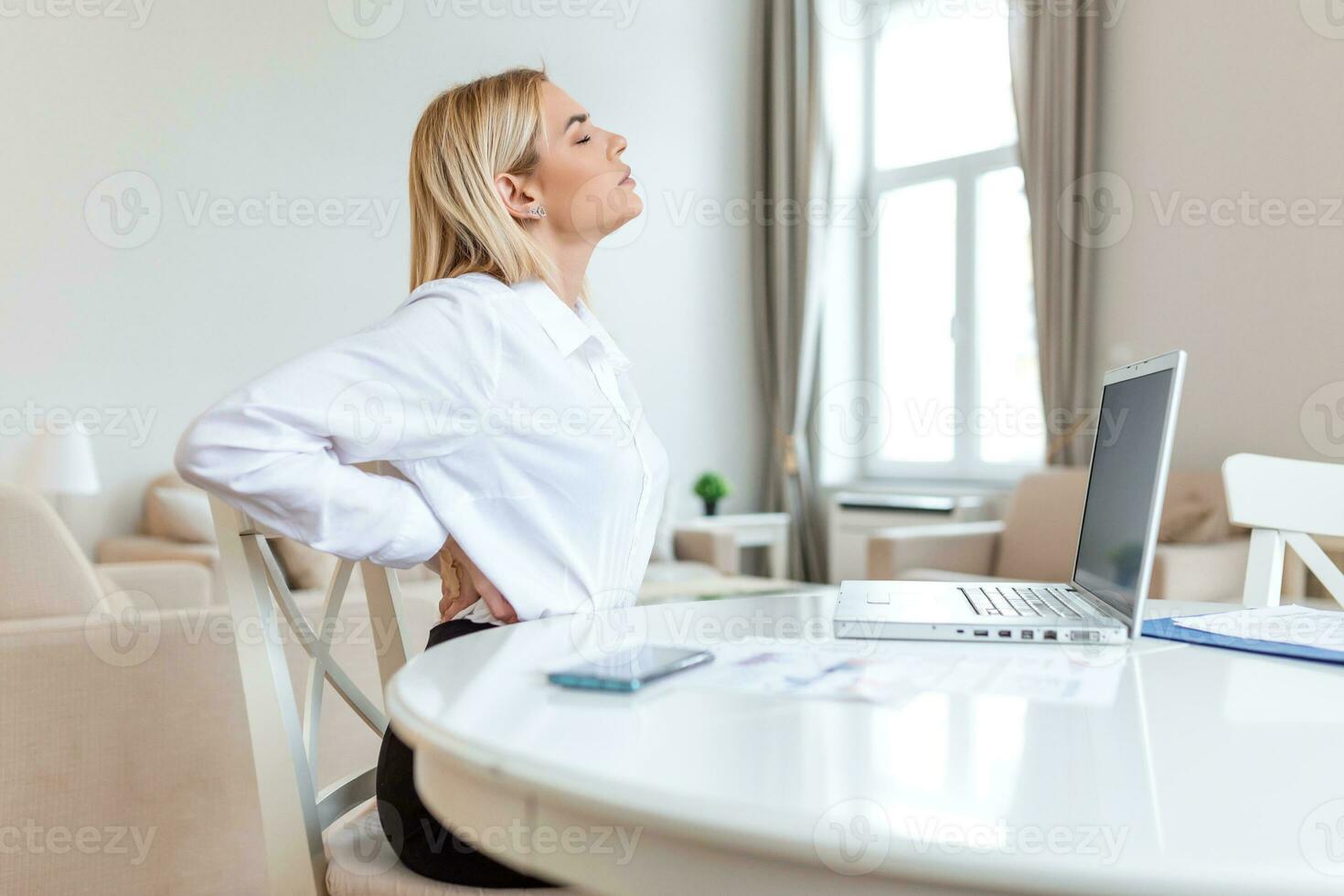 portret van jong benadrukt vrouw zittend Bij huis kantoor bureau in voorkant van laptop, aanraken pijn doen terug met gepijnigd uitdrukking, lijden van rugpijn na werken Aan pc foto